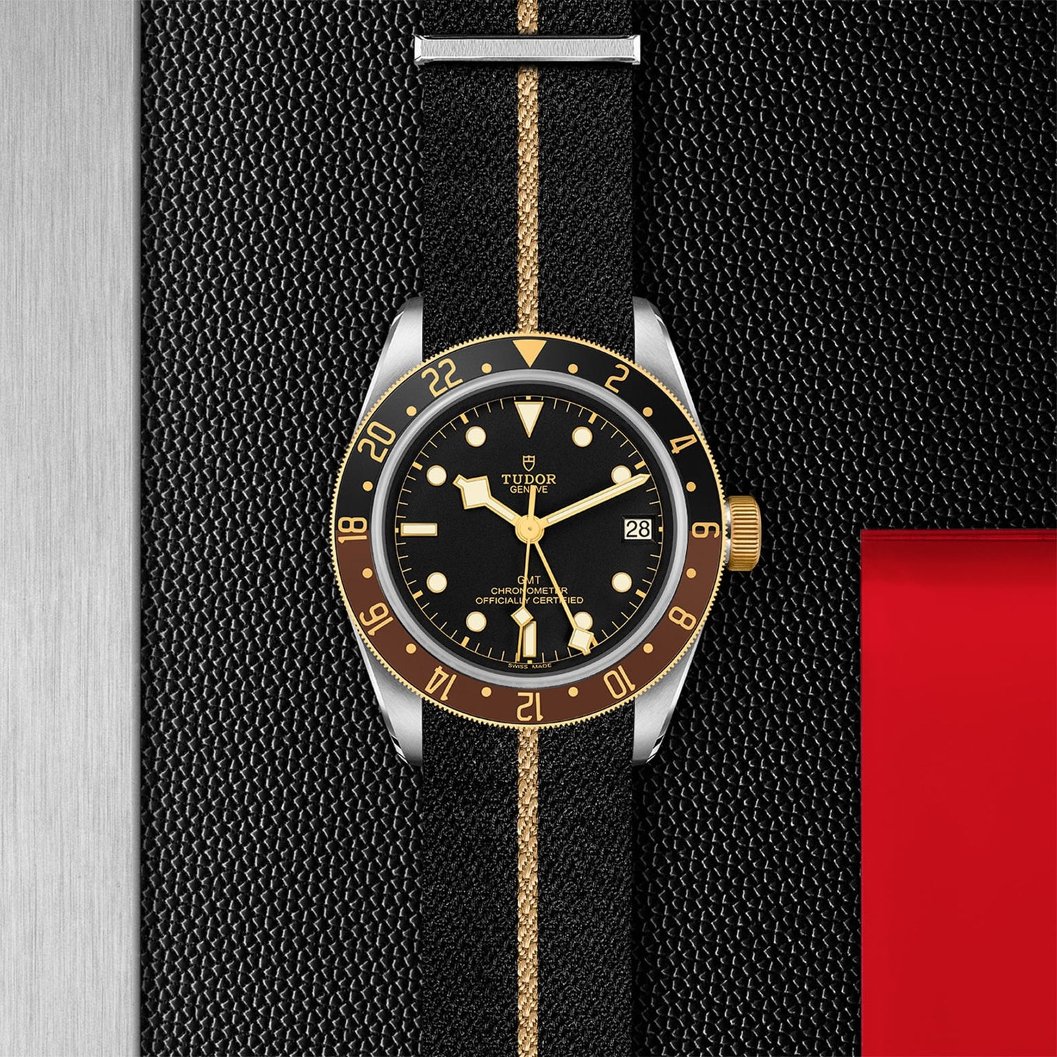 The TUDOR Black Bay GMT S&G, featuring a black and gold bezel, black dial, and a black fabric strap with a gold stripe, is displayed on a textured black and red background, highlighting the elegance of TUDOR diver’s watches.