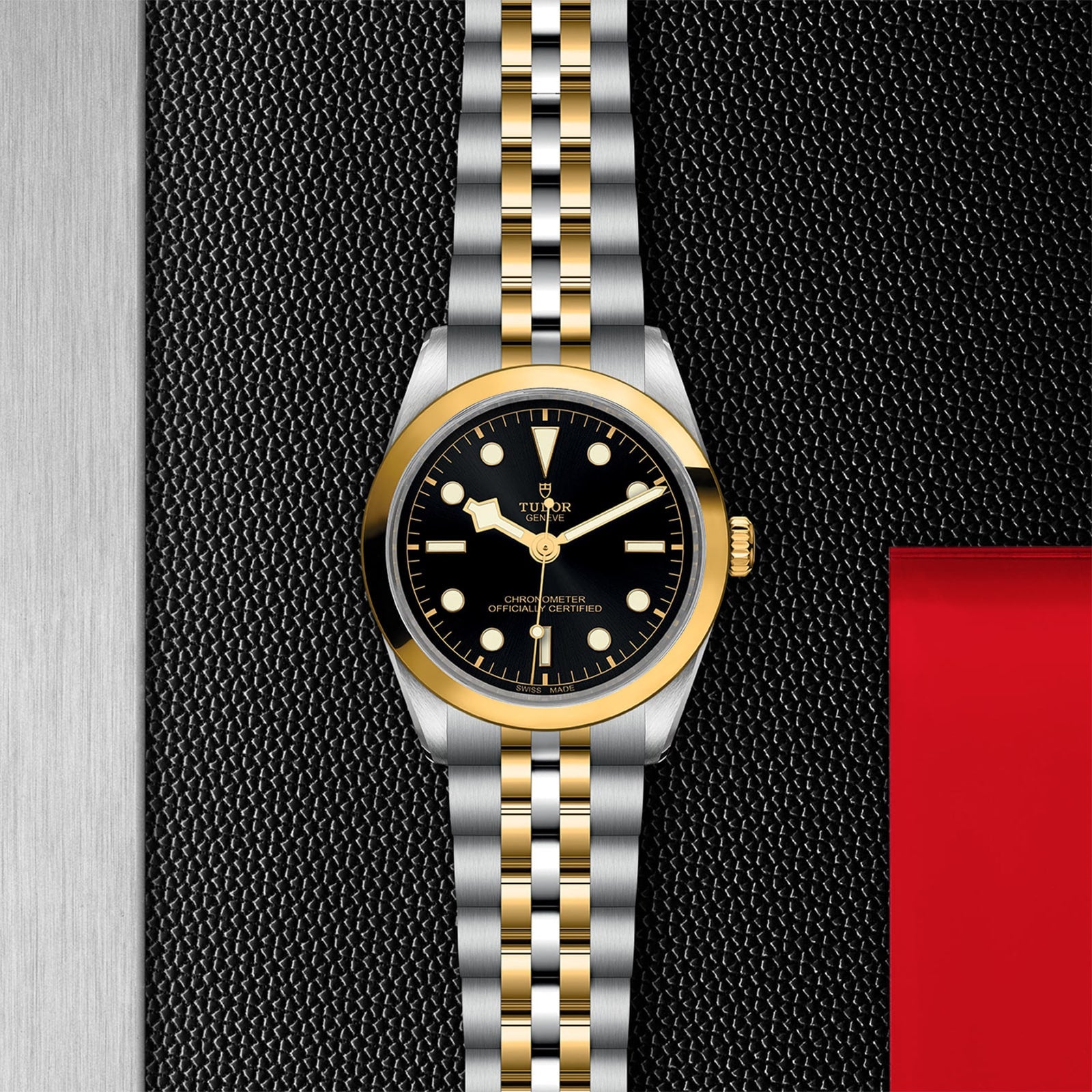 The TUDOR Black Bay One 36 S&G, featuring a black dial with gold accents, is displayed on a textured black and metallic surface beside a red rectangular area.