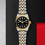 The TUDOR Black Bay One 36 S&G, featuring a black dial with gold accents, is displayed on a textured black and metallic surface beside a red rectangular area.