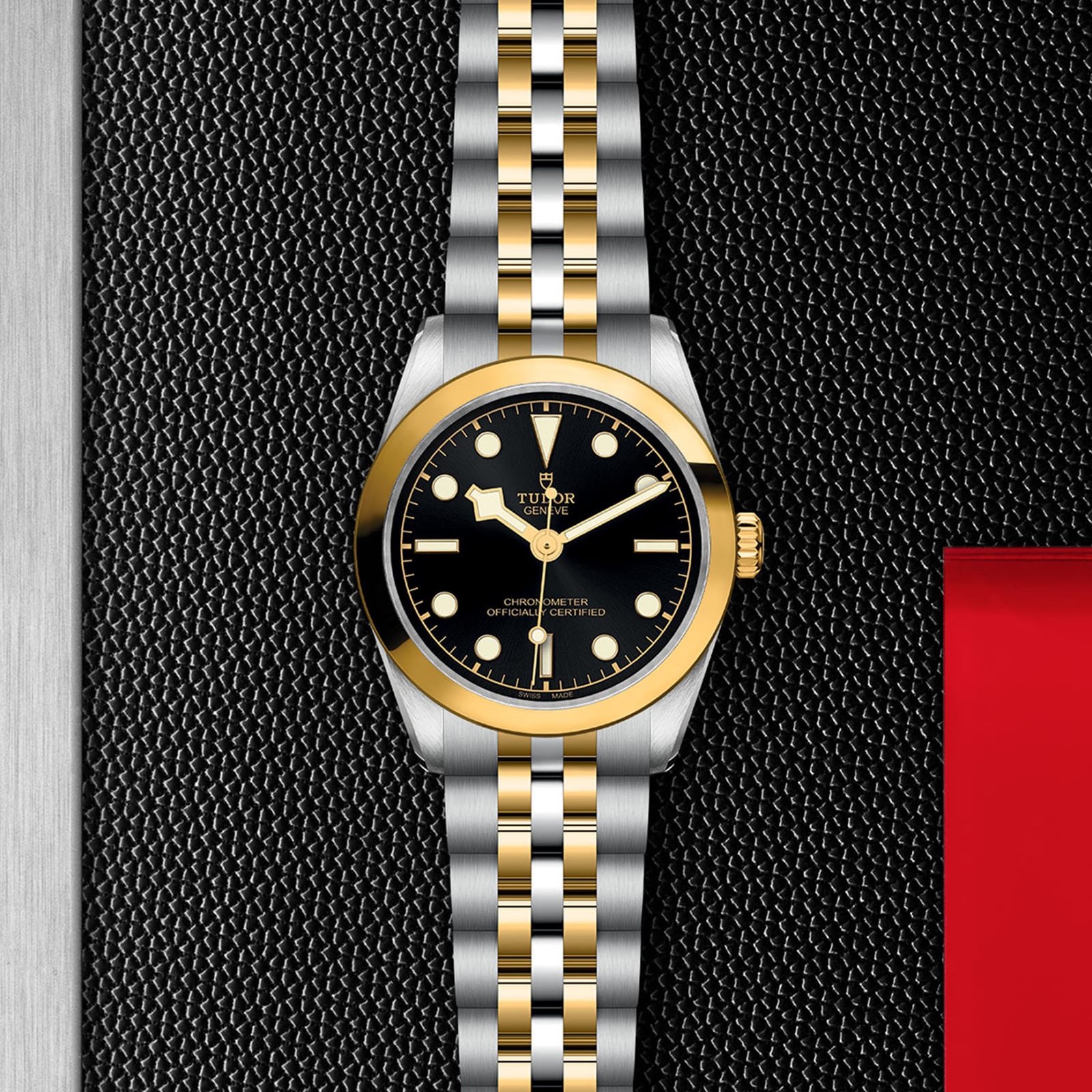 The TUDOR Black Bay One 31 S&G wristwatch, with its black dial, gold bezel, and two-tone gold and silver bracelet, is displayed on a textured black surface, highlighting the iconic TUDOR style and diving watch heritage.