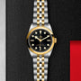 The TUDOR Black Bay One 31 S&G wristwatch, with its black dial, gold bezel, and two-tone gold and silver bracelet, is displayed on a textured black surface, highlighting the iconic TUDOR style and diving watch heritage.