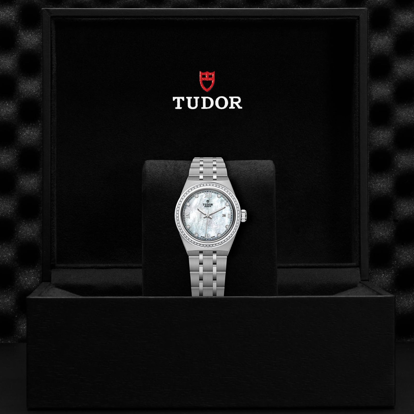A TUDOR Royal wristwatch with a silver metal band and white face is displayed in an open black TUDOR-branded box.