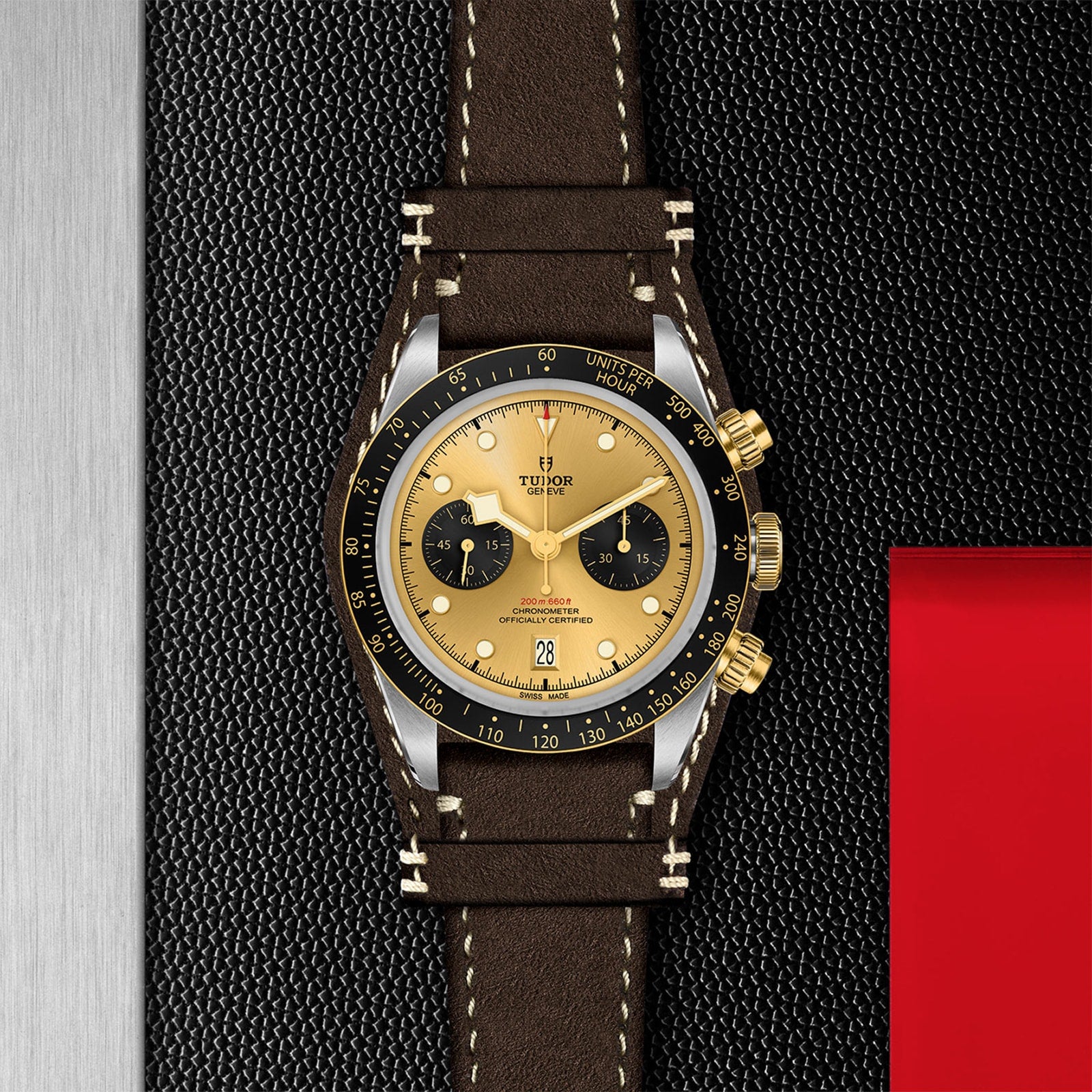 The TUDOR Black Bay Chrono S&G, a gold and black chronograph with a brown leather strap, is shown on a textured black surface beside a metal panel and a red rectangular object.