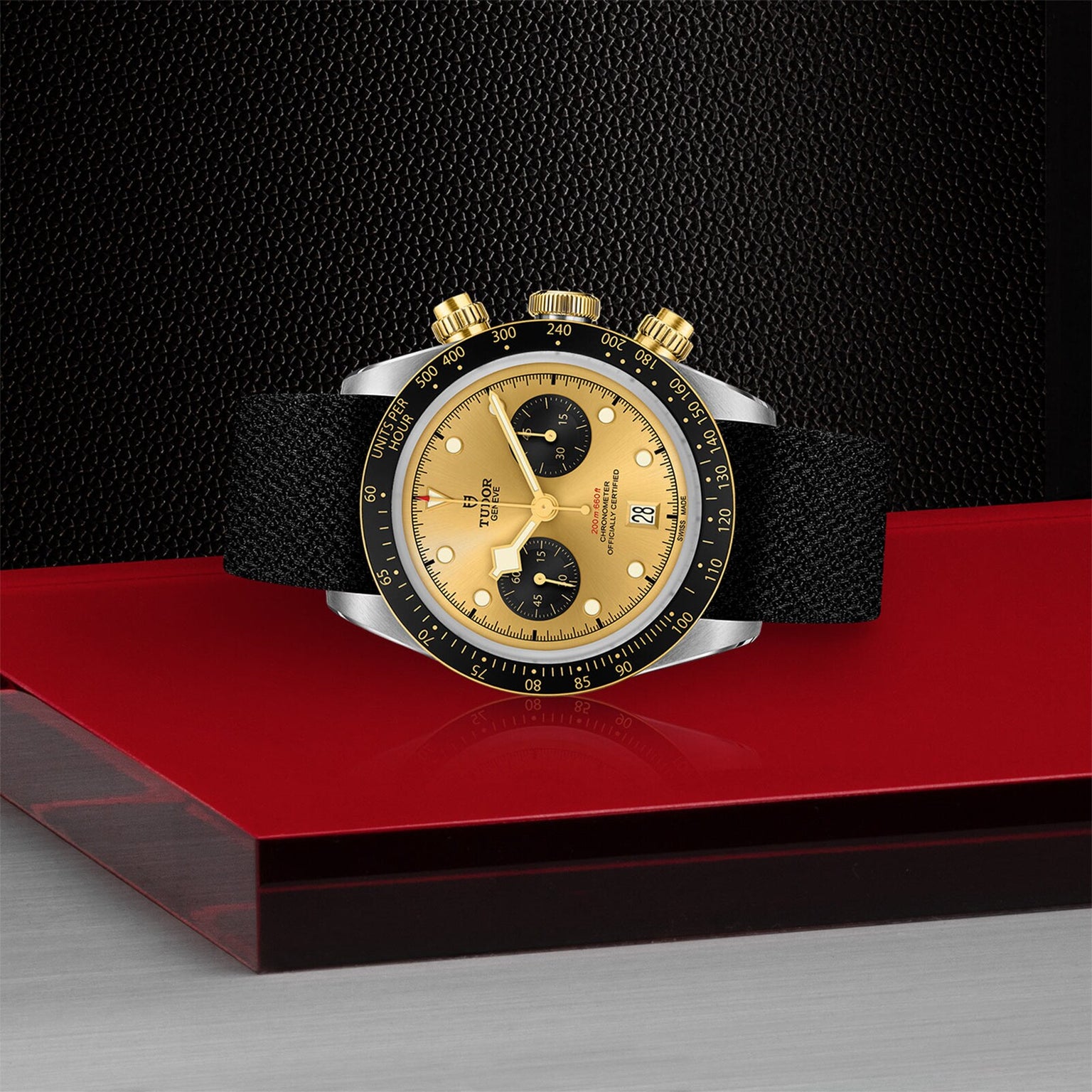 The TUDOR Black Bay Chrono S&G, a gold and black wristwatch with two subdials, black bezel, and black strap, is displayed on a red and gray surface against a black textured background.