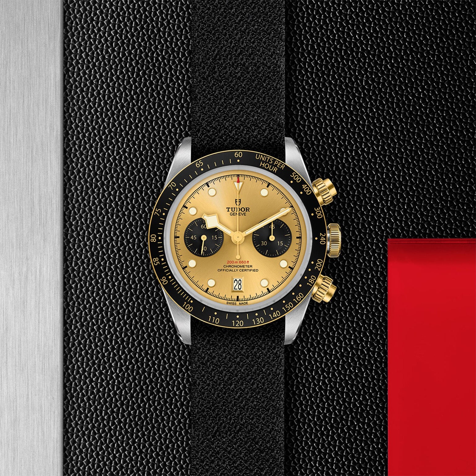 A TUDOR Black Bay Chrono S&G wristwatch featuring a gold dial, black subdials, and a black leather strap is displayed on a textured black background with metallic and red accents.