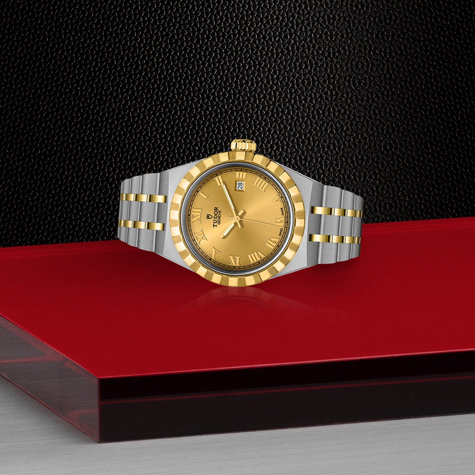 A TUDOR Royal wristwatch in two-tone gold and stainless steel, featuring a gold dial with Roman numerals, is displayed on a red and black geometric surface against a textured black background.