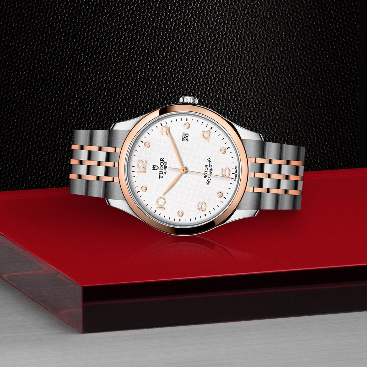 A TUDOR 1926 wristwatch with a silver and rose gold band, showcased against a striking red and black background.