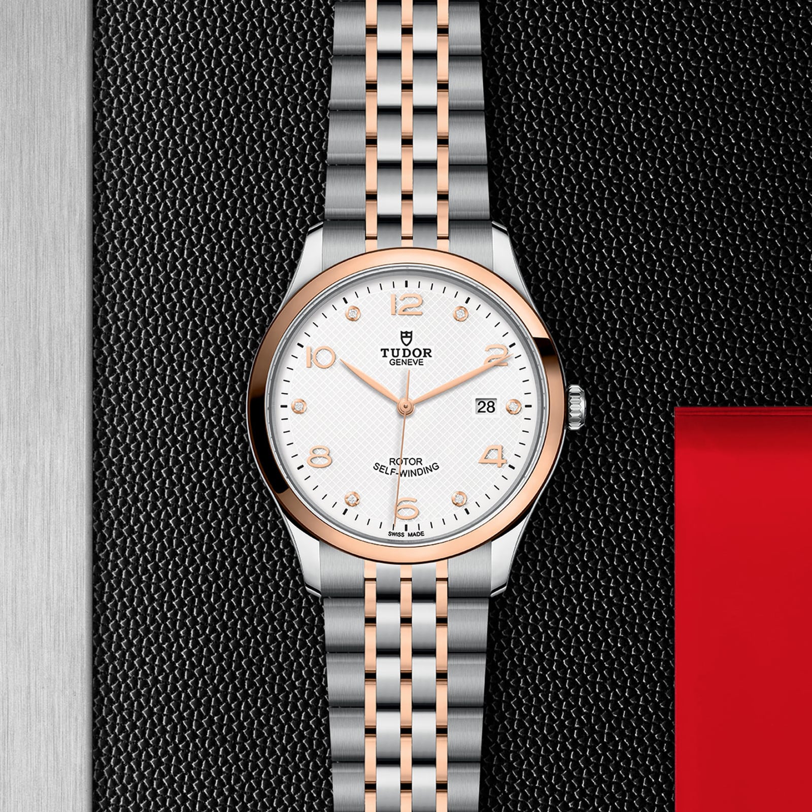 A TUDOR 1926 wristwatch with a silver and rose gold band, white dial, gold hour markers, and date display rests on a black textured surface—crafted in the tradition of mechanical watches by Rolex founder Hans Wilsdorf.
