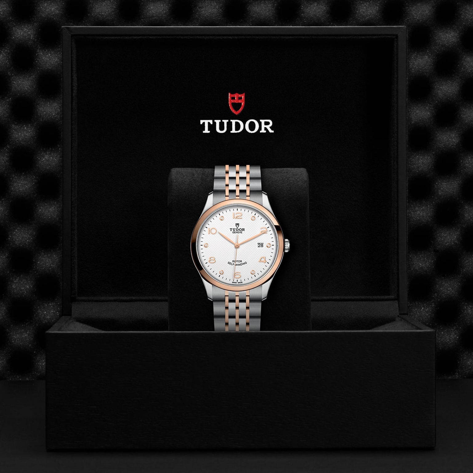 A TUDOR 1926 wristwatch with a two-tone metal band and white dial is displayed in an open black TUDOR-branded box.