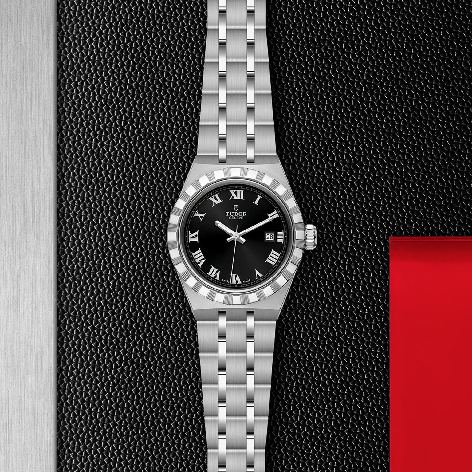 A TUDOR Royal wristwatch with a black dial, Roman numerals, and date display sits on a textured black surface with metal and red accents—showcasing the elegance of TUDOR’s automatic sport-chic design.