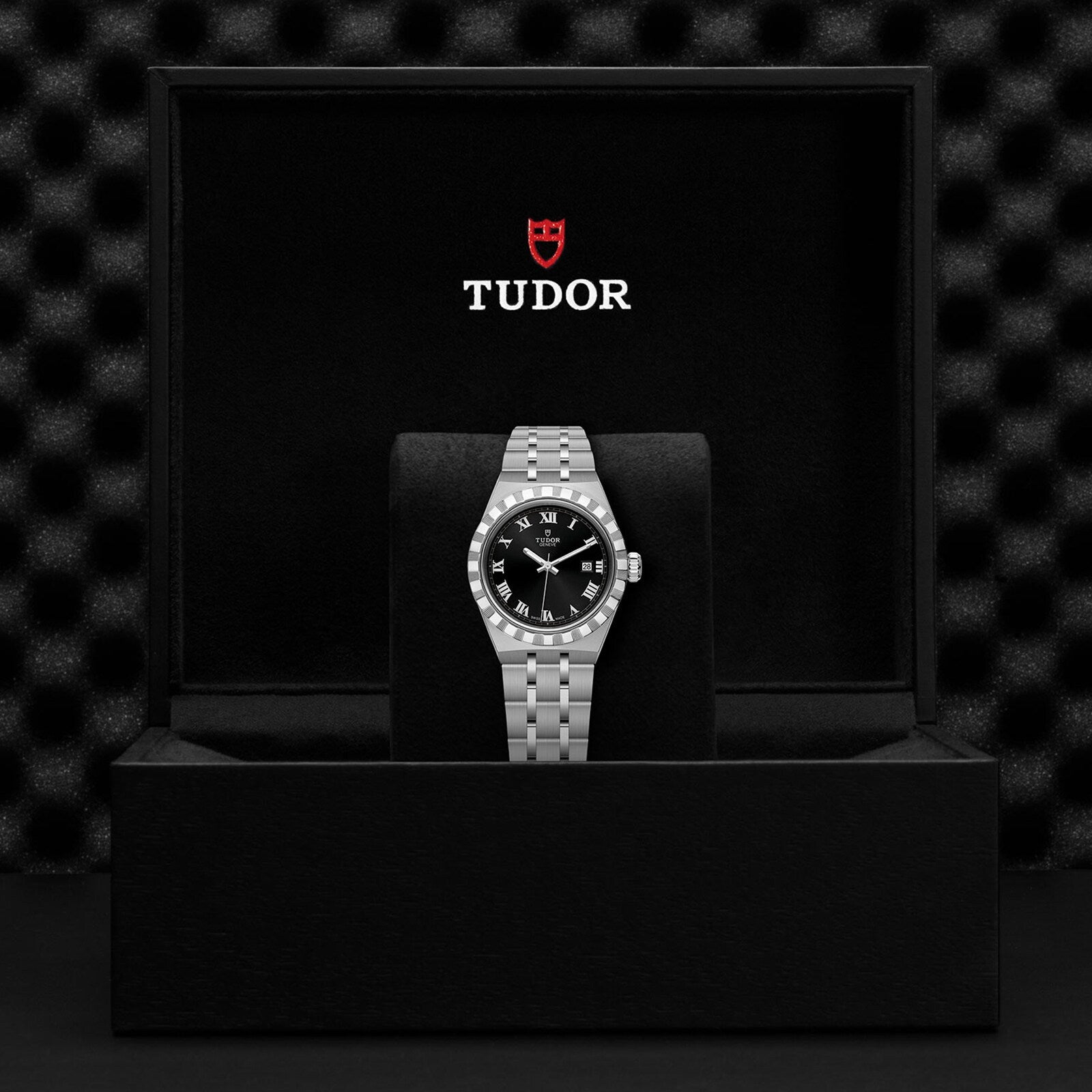 A TUDOR Royal wristwatch, featuring a black dial with Roman numerals, is presented on a cushion inside a black TUDOR box, highlighting the sport-chic elegance of this automatic stainless steel and gold timepiece.