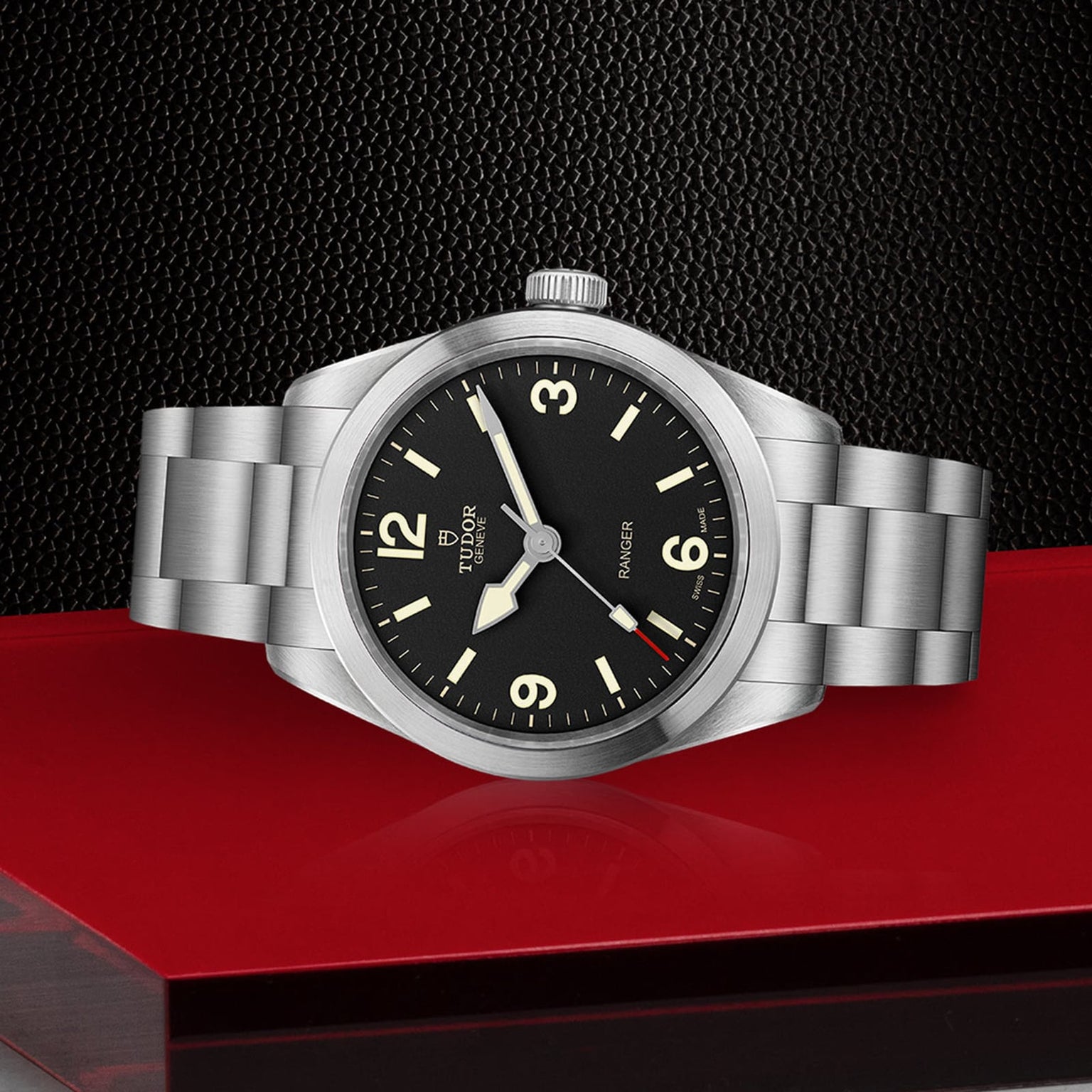 A TUDOR Ranger with a black dial, Arabic numerals, and Super Luminova markers sits on a stainless steel bracelet, showcased against a textured black and red backdrop.