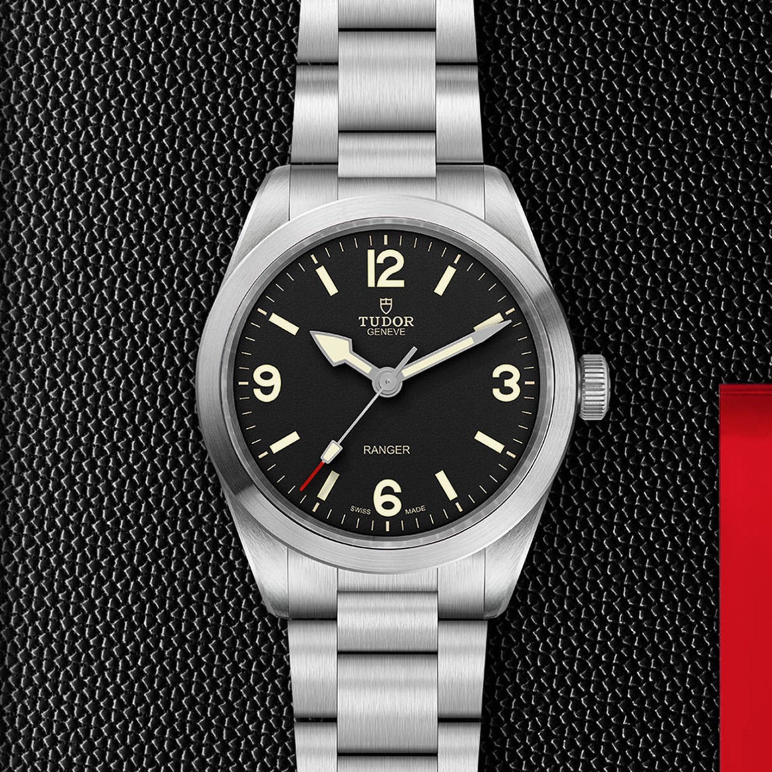 A TUDOR Ranger watch with a black dial, white Super Luminova markers, and a stainless steel bracelet is shown on a textured black surface featuring a red accent on the right.