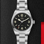 A TUDOR Ranger watch with a black dial, white Super Luminova markers, and a stainless steel bracelet is shown on a textured black surface featuring a red accent on the right.