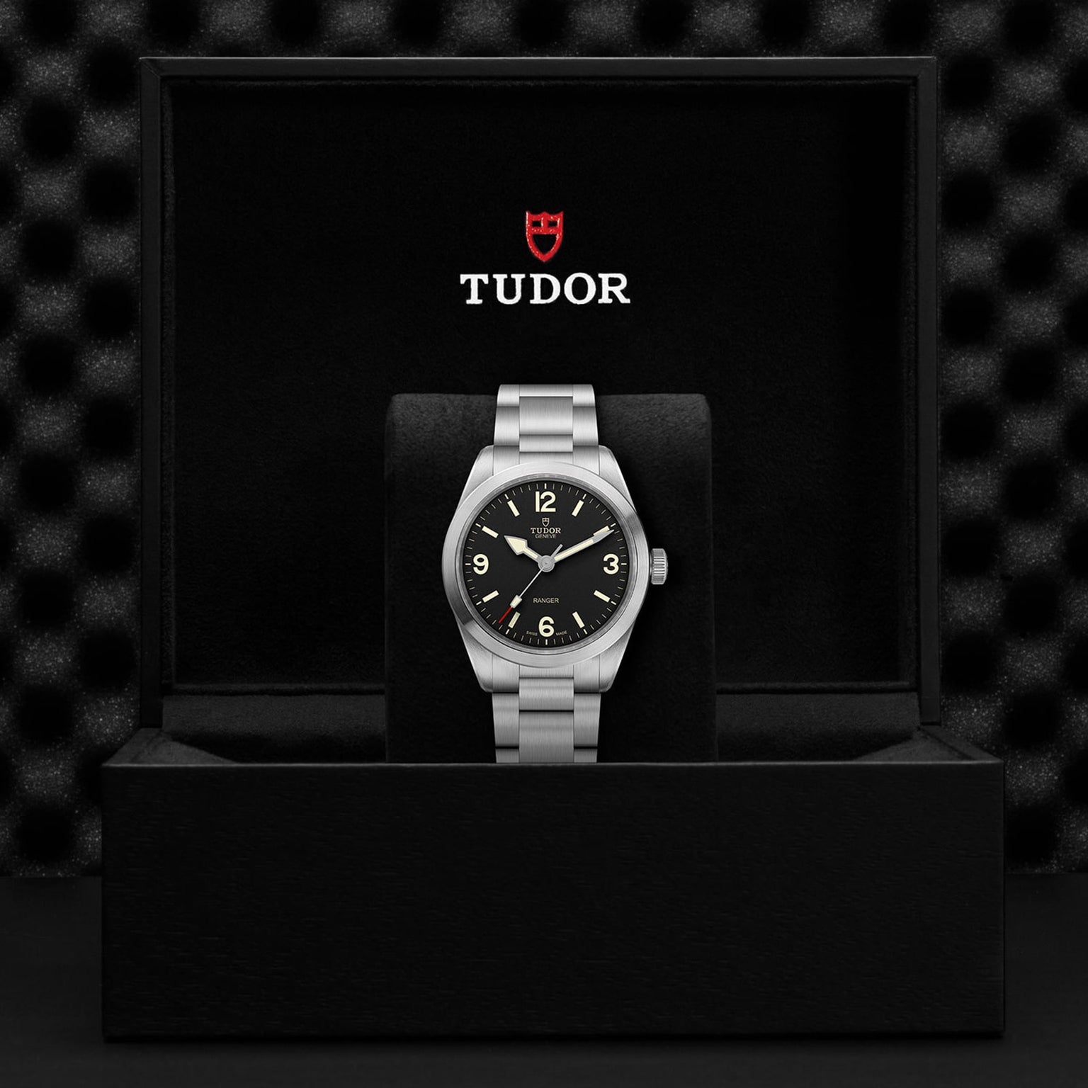 A TUDOR Ranger watch with a black dial and Super Luminova markers is shown upright in an open black box with the TUDOR logo on the inside lid.
