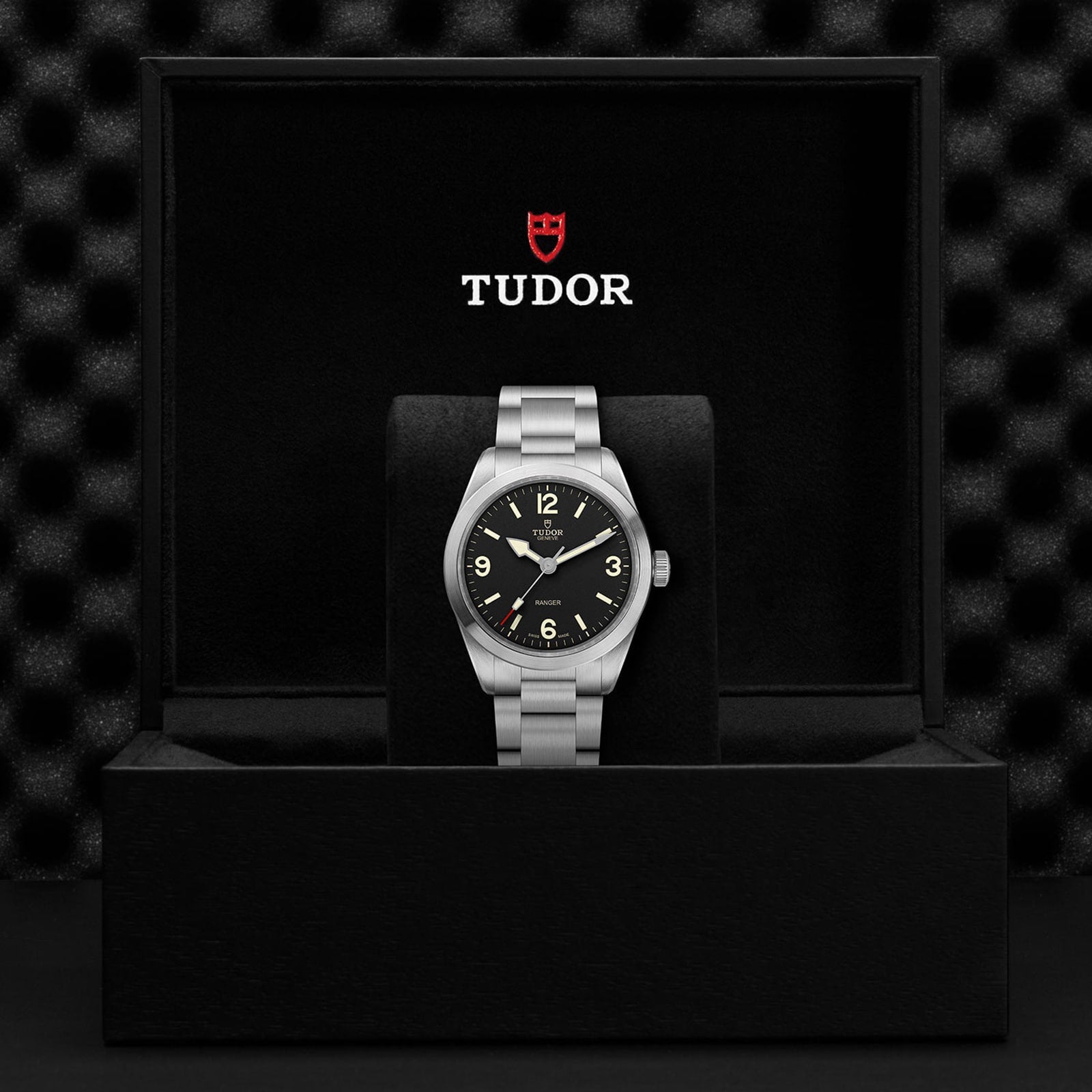 A TUDOR Ranger watch with a black dial and Super Luminova markers is shown upright in an open black box with the TUDOR logo on the inside lid.