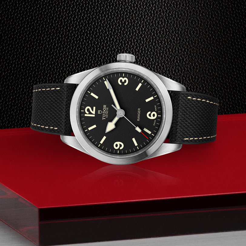 The TUDOR Ranger wristwatch, featuring Super Luminova on its black dial, silver bezel, and black textured strap, is displayed against a bold black and red geometric backdrop.