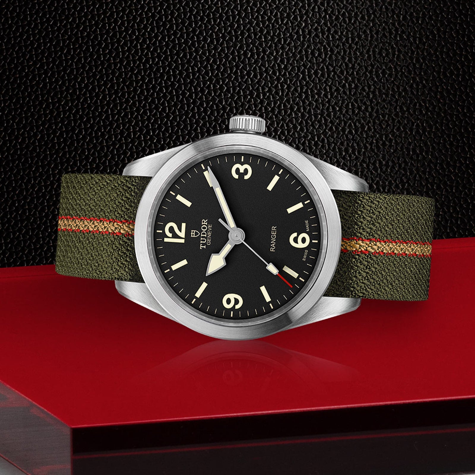 The TUDOR Ranger wristwatch, featuring a black dial with white numerals and an olive green jacquard strap, is shown on a red surface against a textured black background. It’s powered by the TUDOR MT5402 movement.