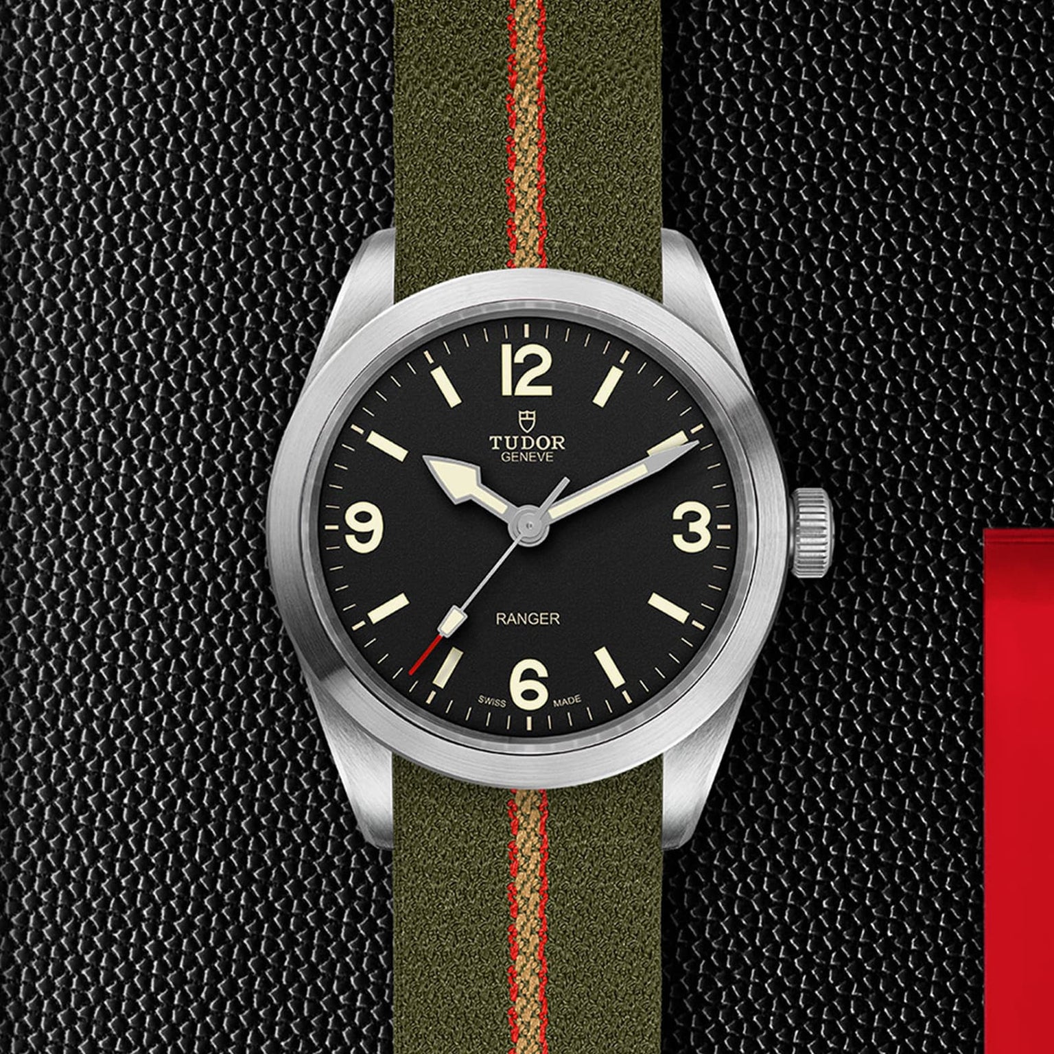 The TUDOR Ranger wristwatch features a black dial with white numerals and an olive green jacquard weave strap with a red stripe, shown on a textured black surface.
