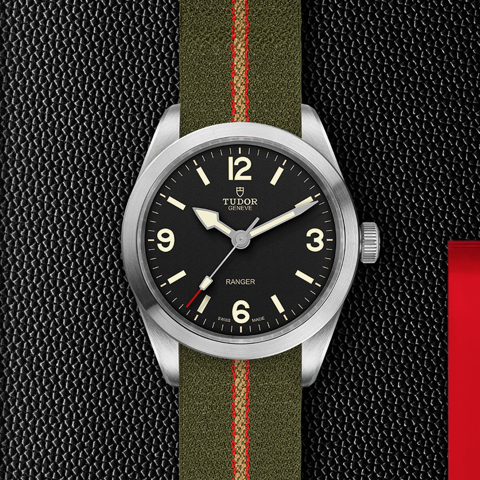 The TUDOR Ranger wristwatch features a black dial with white numerals and an olive green jacquard weave strap with a red stripe, shown on a textured black surface.