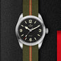 The TUDOR Ranger wristwatch features a black dial with white numerals and an olive green jacquard weave strap with a red stripe, shown on a textured black surface.