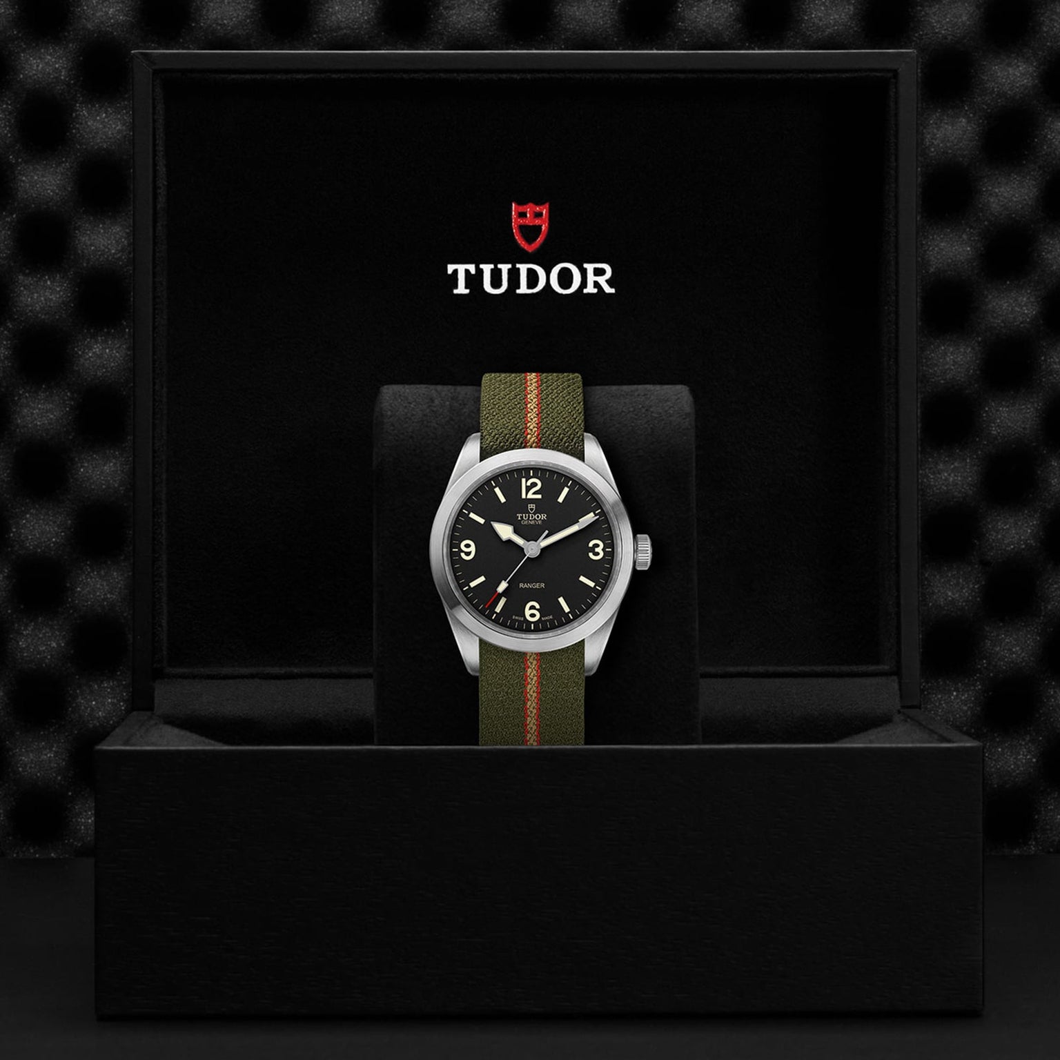 A TUDOR Ranger wristwatch with a black dial and an olive green jacquard strap is showcased upright in a black watch box displaying the TUDOR logo on the underside of the lid.