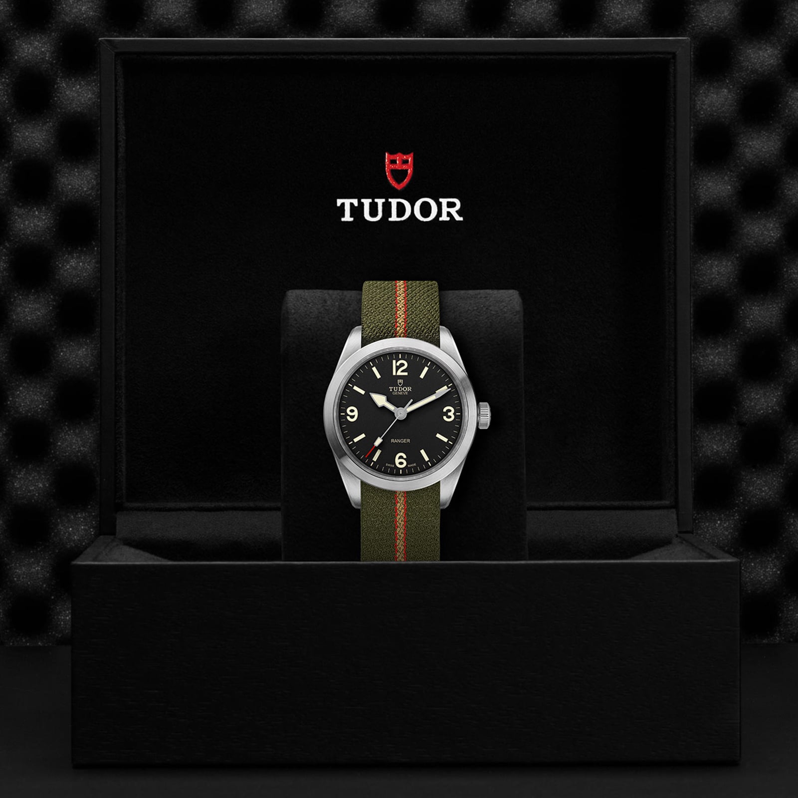 A TUDOR Ranger wristwatch with a black dial and an olive green jacquard strap is showcased upright in a black watch box displaying the TUDOR logo on the underside of the lid.