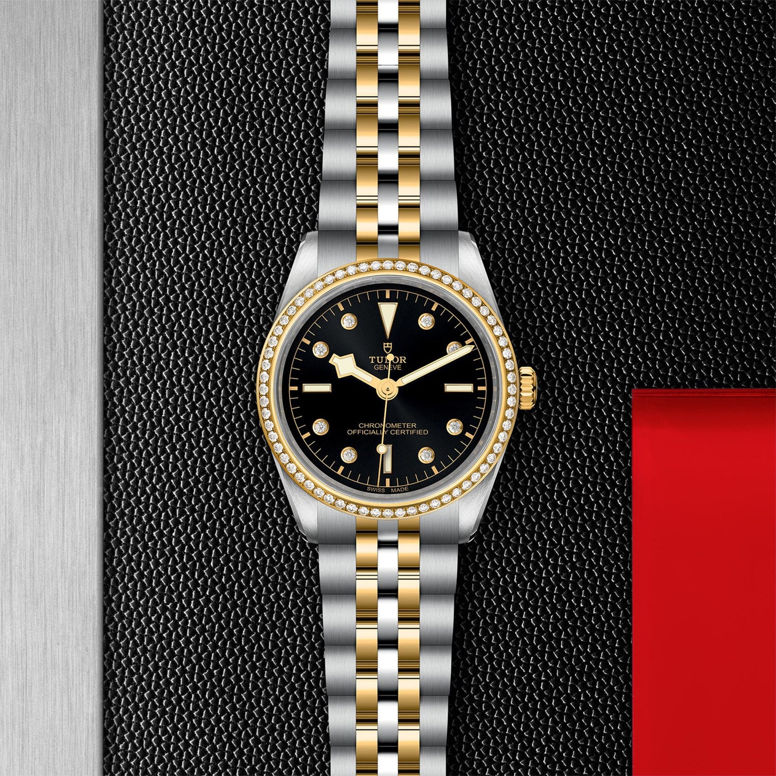 The TUDOR Black Bay One 36 S&G features a black dial with yellow gold hour markers and a two-tone gold and silver bracelet, elegantly displayed on a textured black surface.