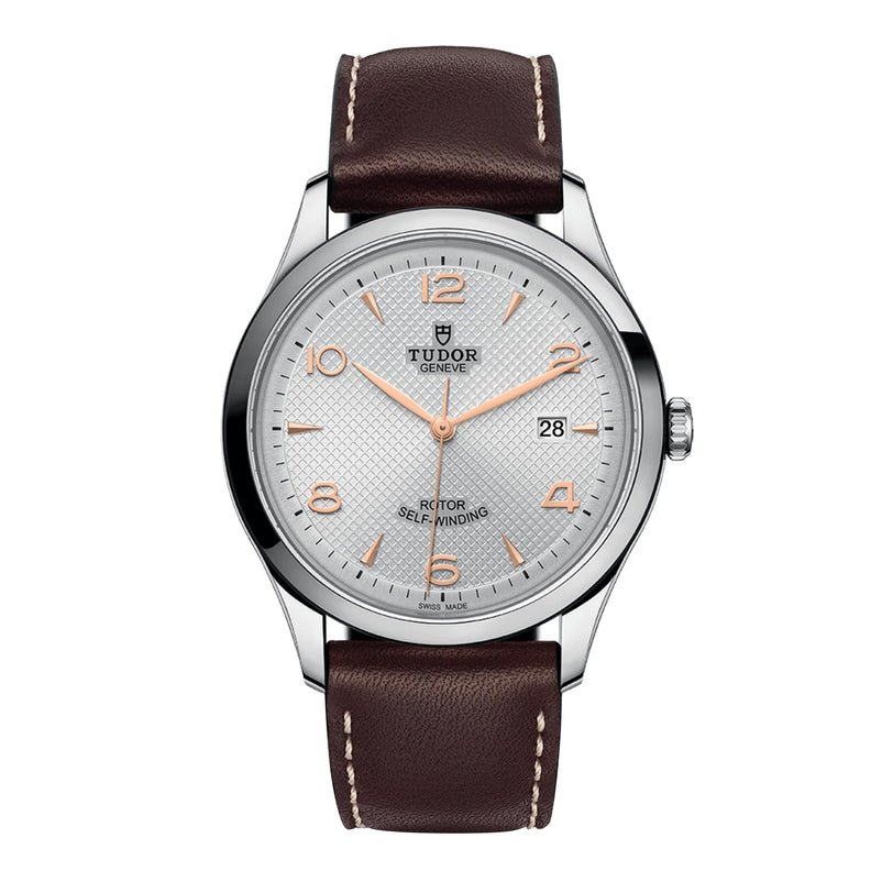 A TUDOR 1926 wristwatch featuring a brown leather strap, silver case, white textured dial, rose gold indices and hands, and a 3 o’clock date window—crafted by the brand founded by Rolex’s creator.