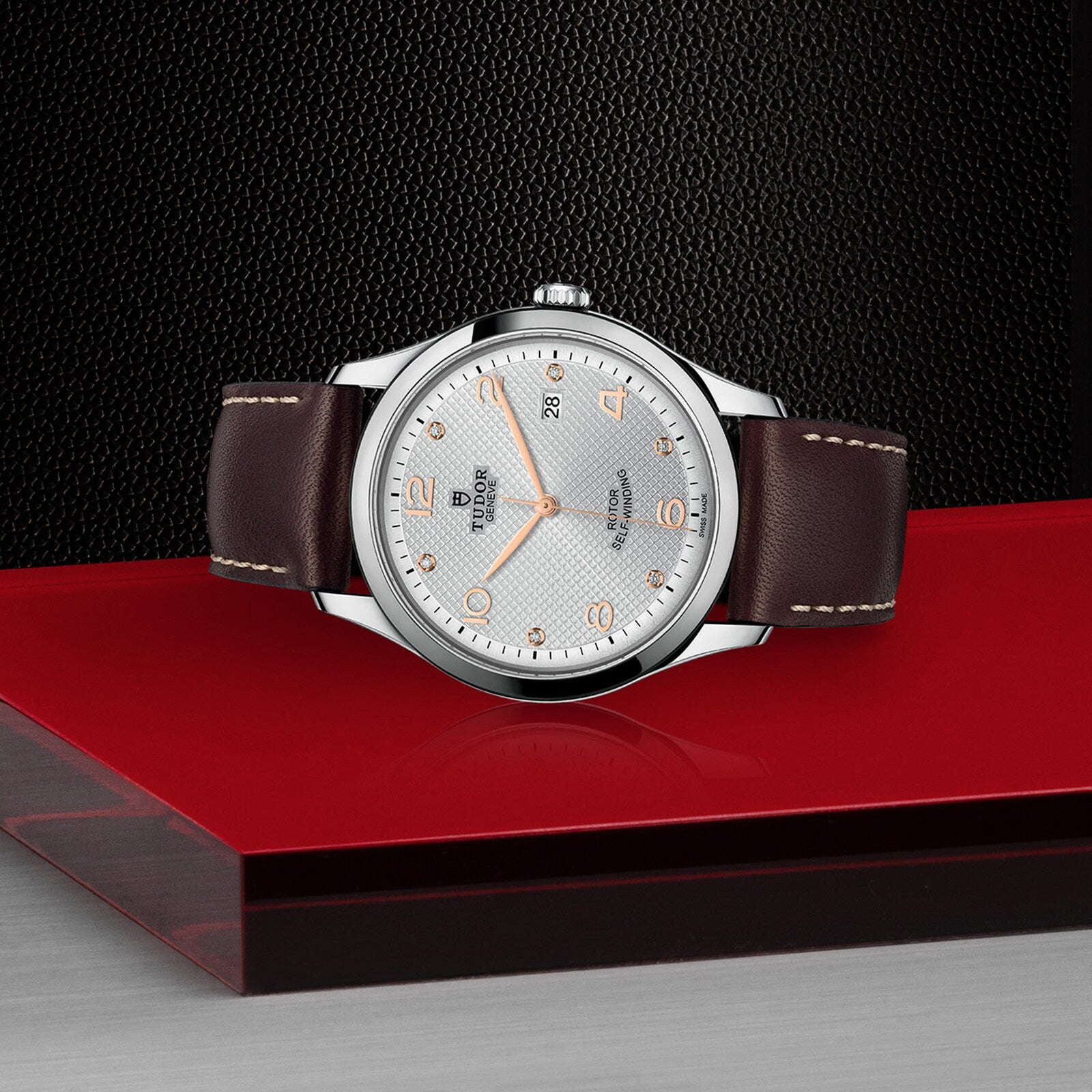 The TUDOR 1926, a classic silver wristwatch with a brown leather strap, features a mechanical date display and silver-gold hour markers. It rests elegantly on a red and black geometric surface.