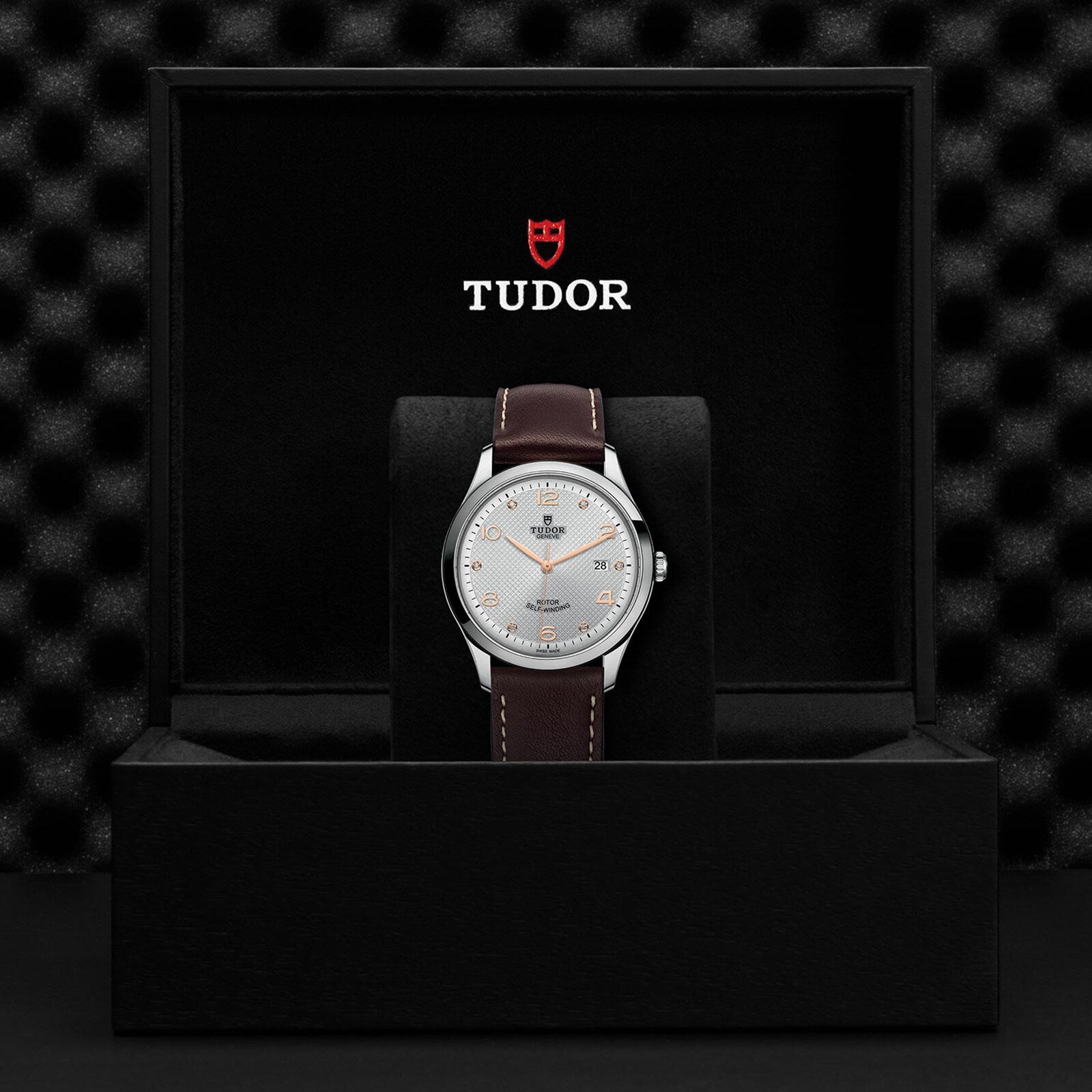 The TUDOR 1926 wristwatch, featuring a silver dial and brown leather strap, is displayed upright in a black box with the Tudor logo inside—a timeless choice for mechanical watch enthusiasts.