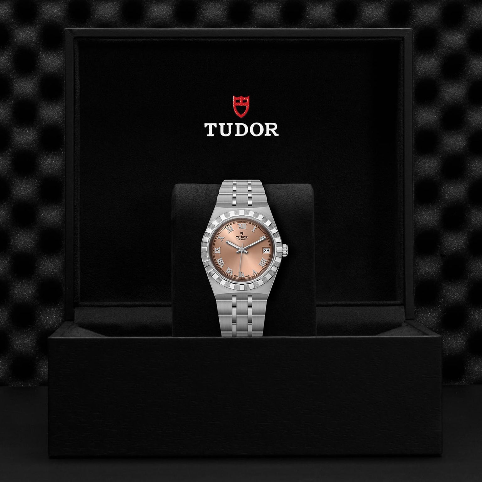 A TUDOR Royal wristwatch with a silver metal band and salmon dial, featuring automatic movement, is displayed upright in a black Tudor-branded presentation box.