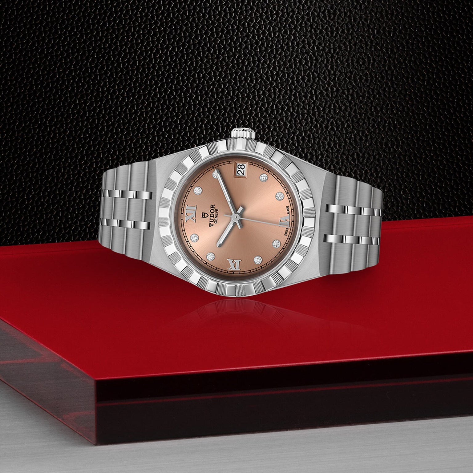 The TUDOR Royal is a silver wristwatch with a rose gold face and diamond markers, shown on a red and black background.