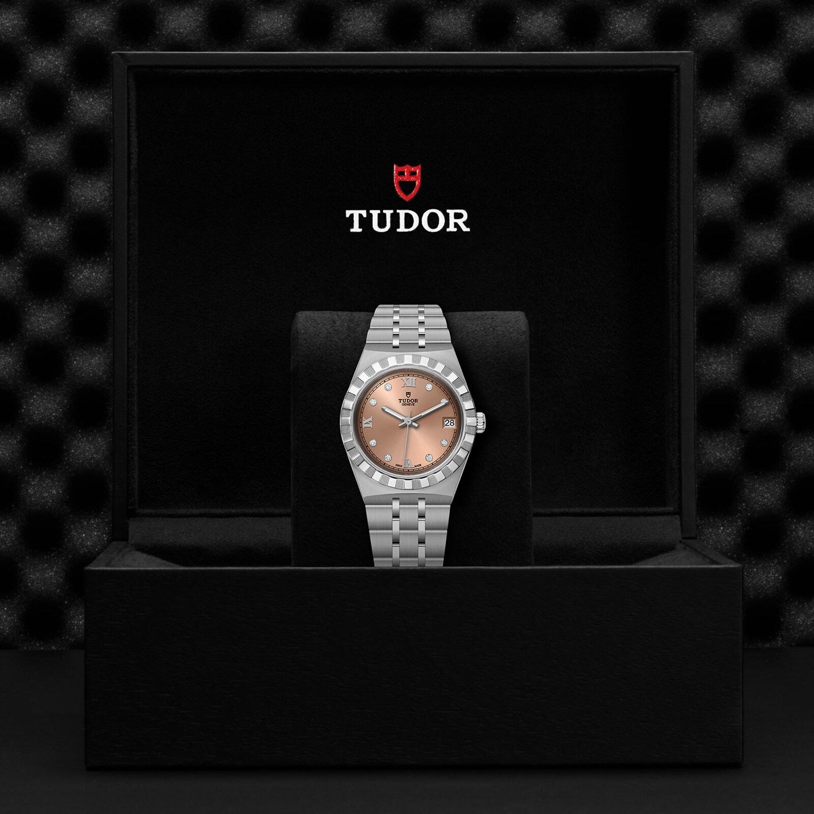 A silver TUDOR Royal wristwatch with a brown dial, date display, and automatic movement is displayed in an open black box featuring the TUDOR logo on the inside lid.
