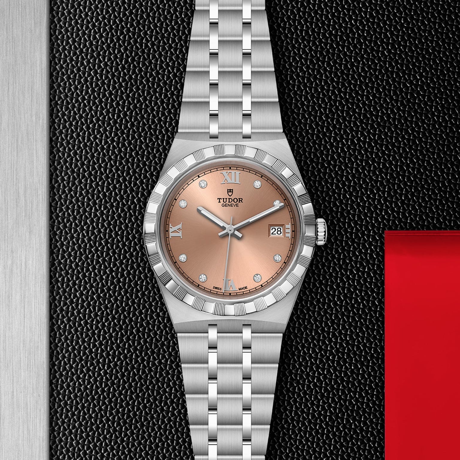 A silver TUDOR Royal wristwatch with a salmon dial, Roman numerals, diamond markers, date display, and automatic movement rests on a textured black surface near a red edge.