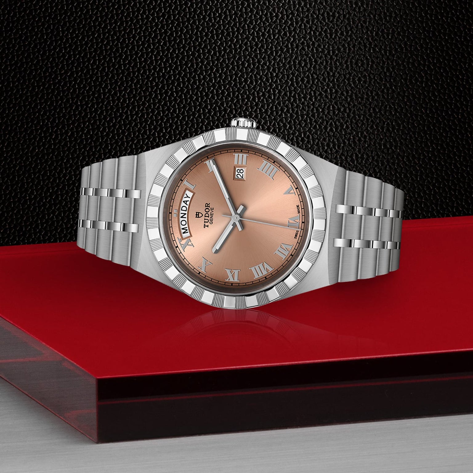 A TUDOR Royal wristwatch with a silver case, salmon dial, Roman numerals, automatic movement, and day-date display is shown on a red and black surface.