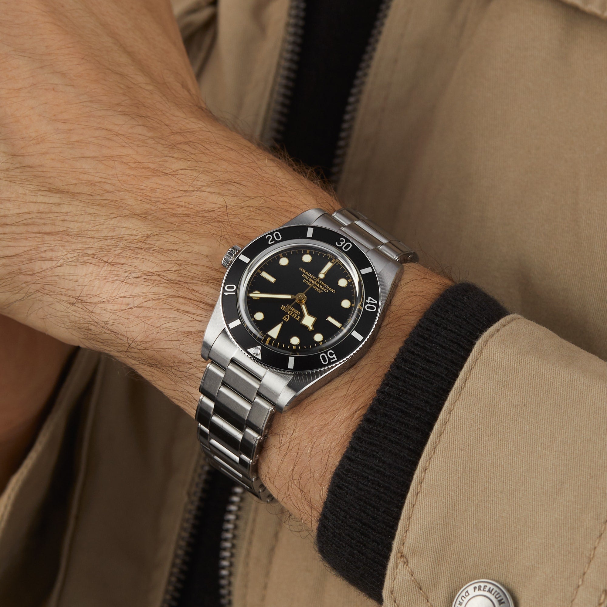 Someone wearing a beige jacket and a TUDOR Black Bay 54 watch with a black dial and bezel, featuring the Manufacture Calibre MT5400, on their wrist.