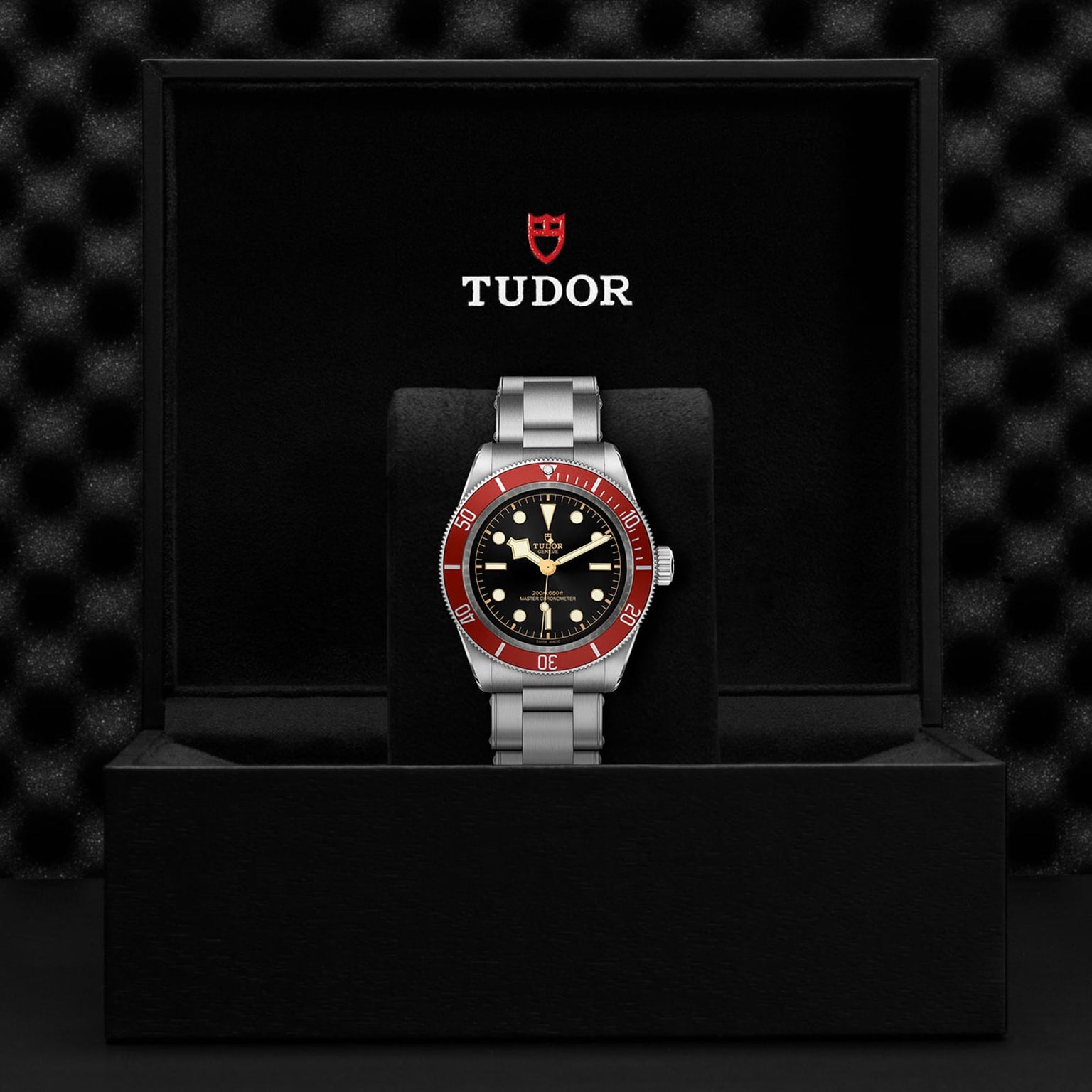 A TUDOR Black Bay wristwatch with a burgundy bezel, black dial, and silver metal band is shown in an open black TUDOR box set against a textured black background.