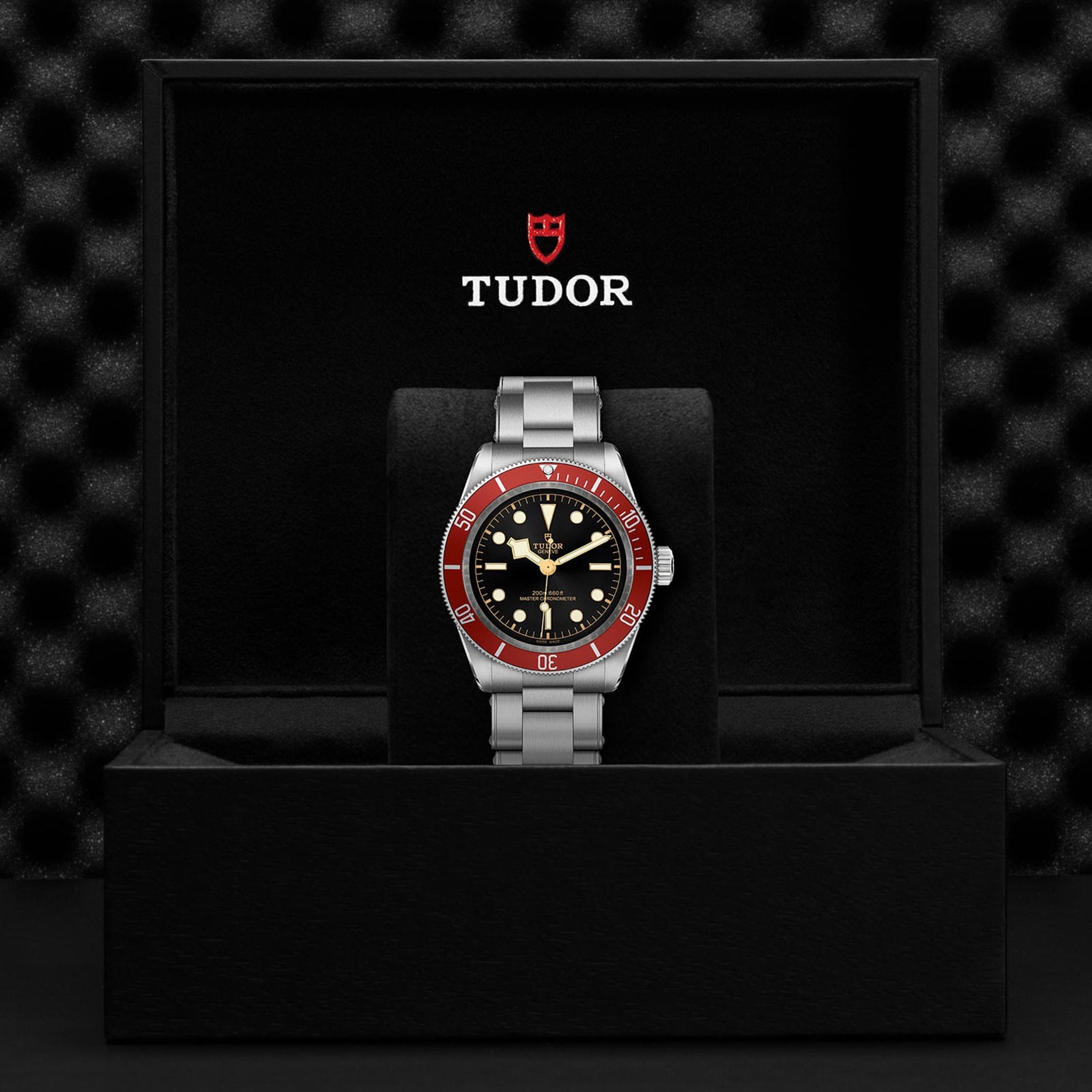 A TUDOR Black Bay wristwatch with a burgundy bezel, black dial, and silver metal band is shown in an open black TUDOR box set against a textured black background.