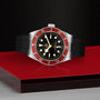 The TUDOR Black Bay wristwatch, featuring a burgundy bezel, black dial, and black rubber strap, is showcased on a red and black textured surface.