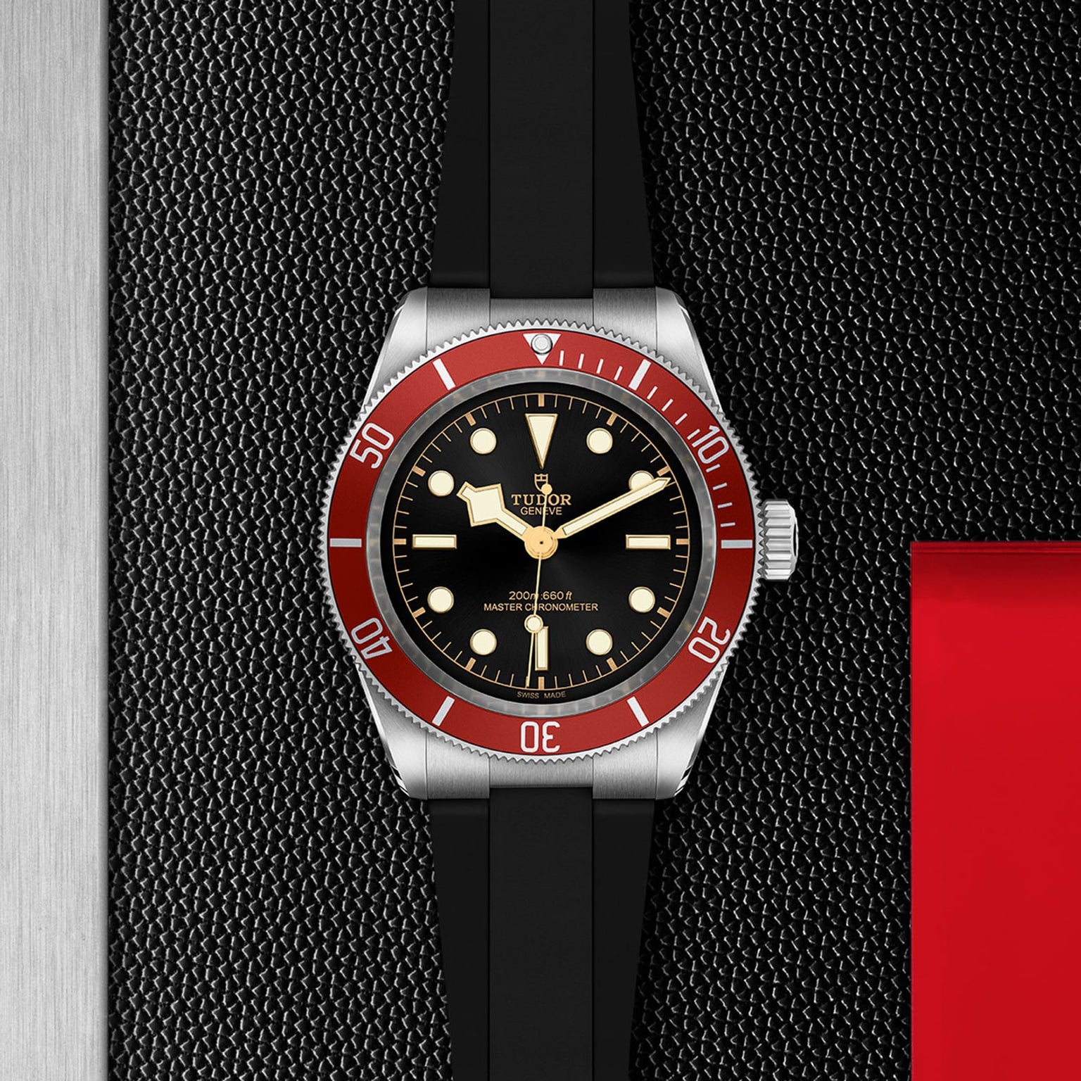 A TUDOR Black Bay watch featuring a burgundy bezel, black dial, and black rubber strap is displayed on a textured black surface with metal and red accents.