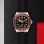 A TUDOR Black Bay watch featuring a burgundy bezel, black dial, and black rubber strap is displayed on a textured black surface with metal and red accents.