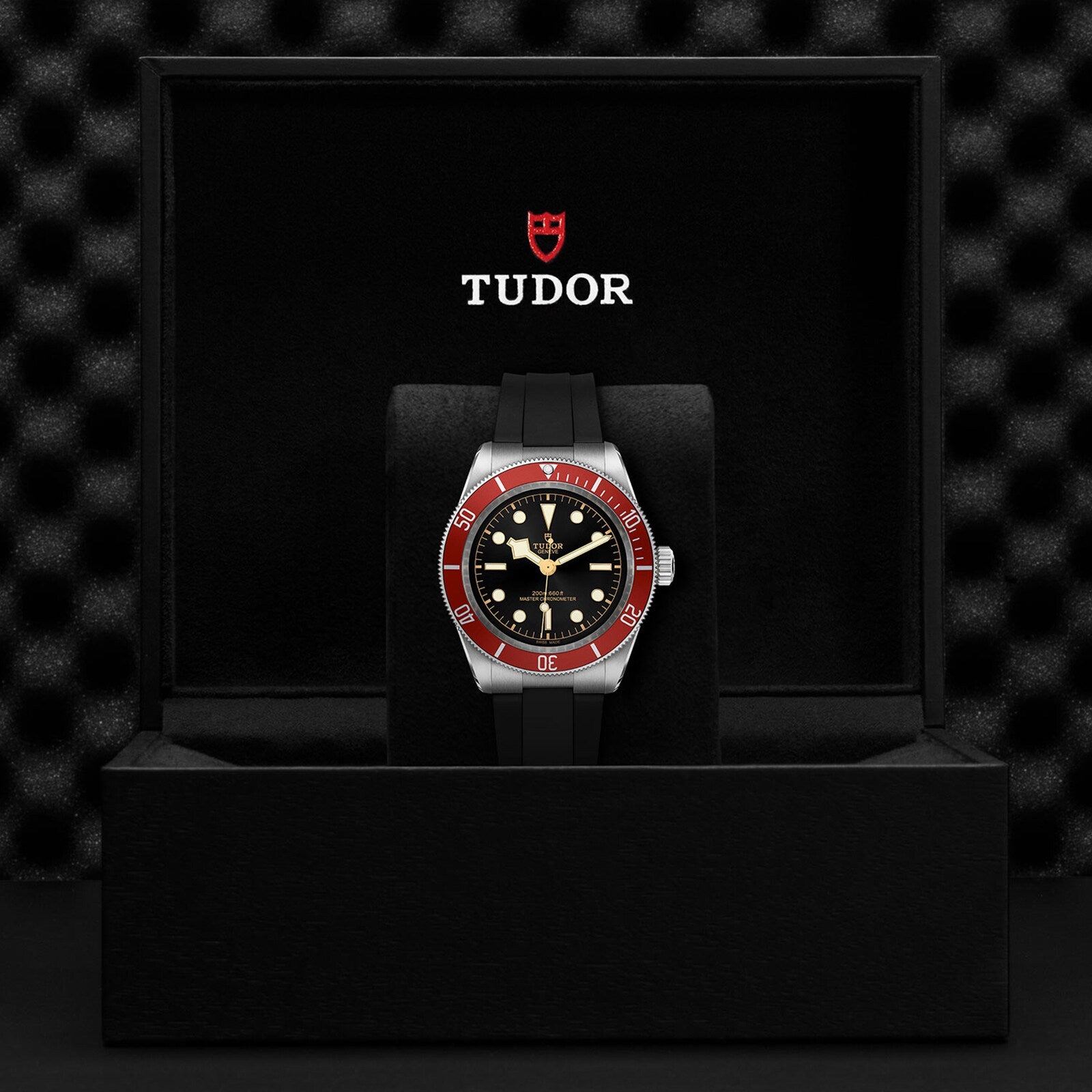 A TUDOR Black Bay wristwatch with a black dial and burgundy bezel on a black strap is displayed in an open black TUDOR box.
