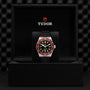 A TUDOR Black Bay wristwatch with a black dial and burgundy bezel on a black strap is displayed in an open black TUDOR box.