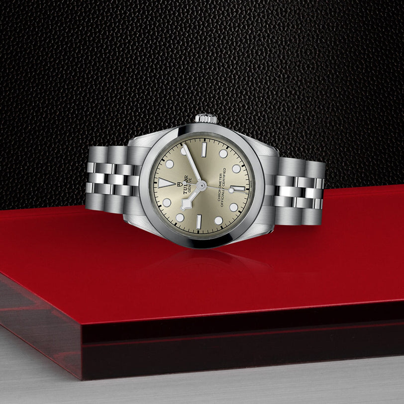 A TUDOR Black Bay One 31 in stainless steel with a champagne dial and bracelet is displayed on a glossy red and black surface, set against a textured black background.