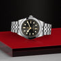 The TUDOR Black Bay One 39, a unisex stainless steel wristwatch with a black dial and silver bezel, is showcased on a glossy red and black surface against a textured black background, capturing the signature elegance of the brand.