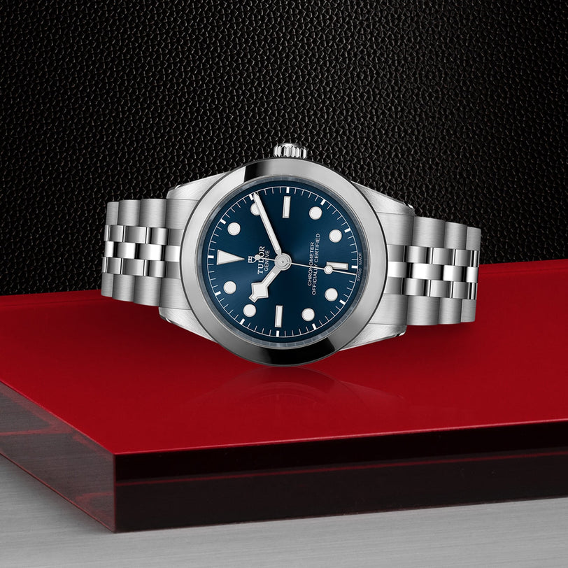 A TUDOR Black Bay One 39 wristwatch in silver with a blue dial, presented on a red and black textured surface.