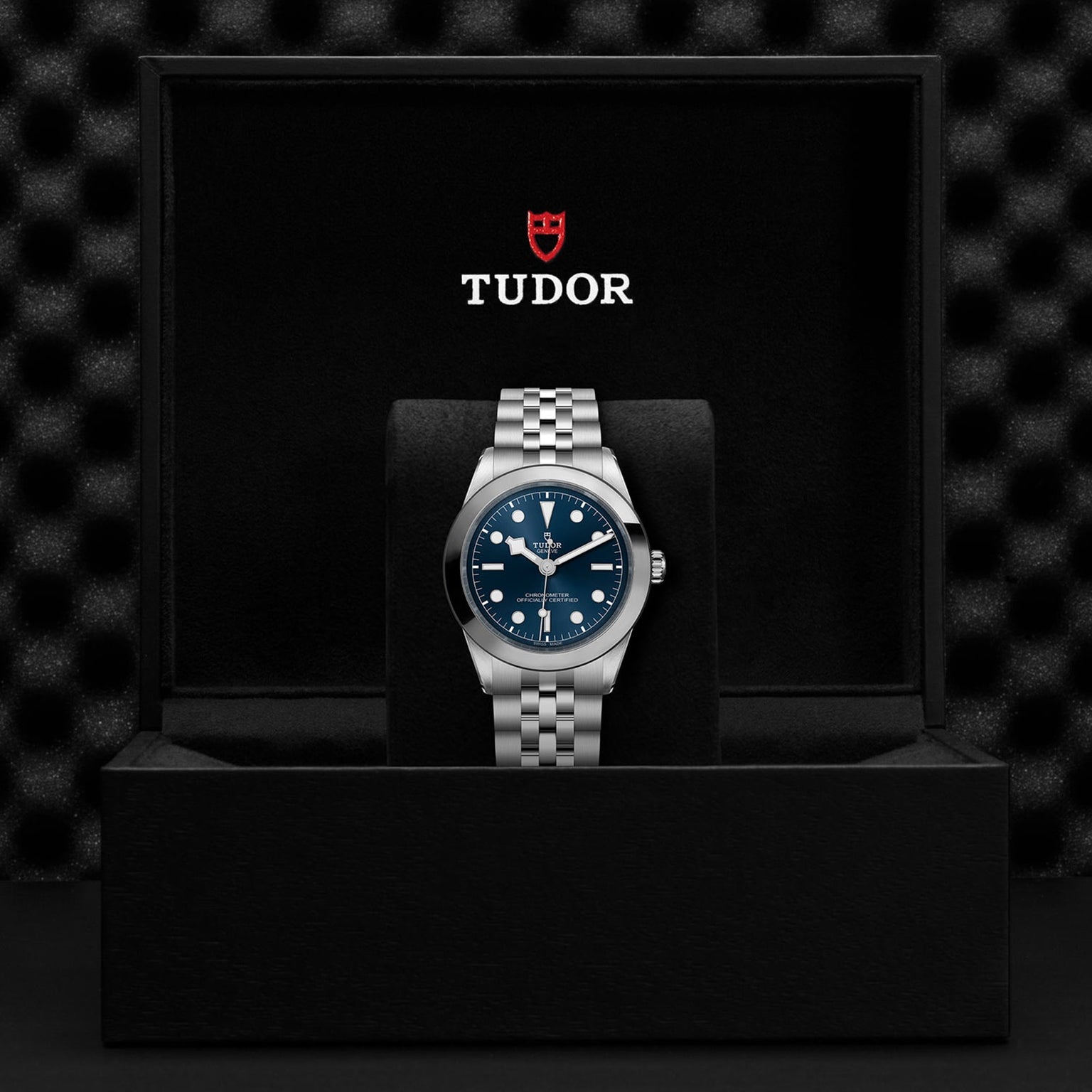 The TUDOR Black Bay One 39, a unisex wristwatch with a blue dial and silver metal band, is showcased upright in its open black box featuring the TUDOR logo, reflecting the refined Black Bay style and advanced Manufacture Calibres.