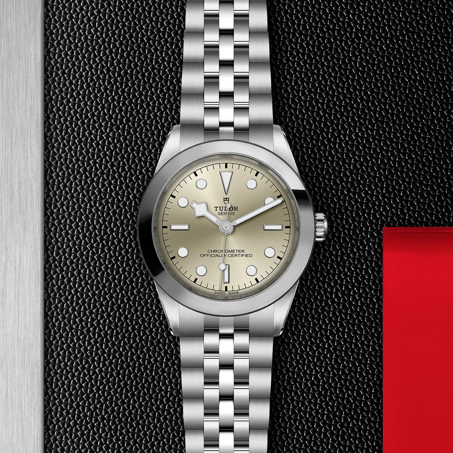 The TUDOR Black Bay One 39 wristwatch, featuring a stainless steel case, five-link silver bracelet, and beige dial, is displayed on a textured black surface, reflecting unisex chic style and lasting elegance.