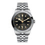 The TUDOR Black Bay One 39 is a unisex stainless steel wristwatch featuring a black dial, gold hour markers, silver bracelet, and date window at 3 o’clock.